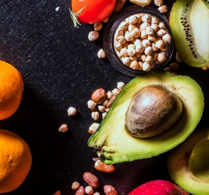 A vibrant arrangement of fruits and nuts on a black background featuring kiwi, avocado, oranges, apple, and peanuts.