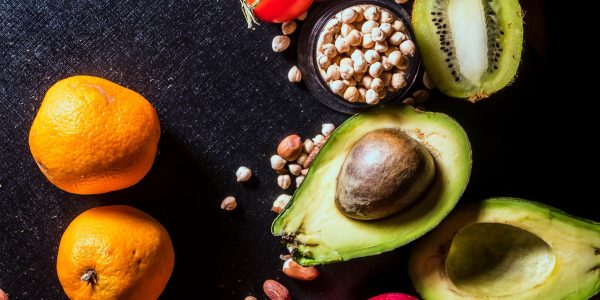 A vibrant arrangement of fruits and nuts on a black background featuring kiwi, avocado, oranges, apple, and peanuts.